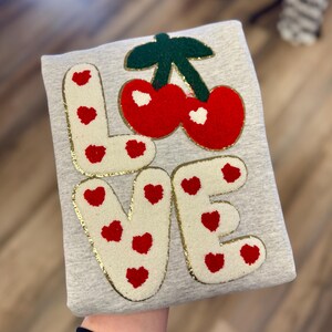 May include: A light gray sweatshirt with the word "LOVE" in white, gold-trimmed letters. The letter shapes are decorated with red hearts. A pair of red cherries with green leaves and gold trim sits above the "VE".