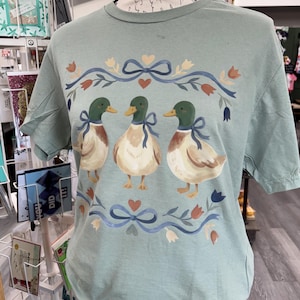 Puede incluir: Una camiseta de manga corta color verde salvia con un estampado gr&aacute;fico de tres patos &aacute;nades reales. Cada pato lleva un lazo azul alrededor del cuello. El dise&ntilde;o est&aacute; enmarcado por lazos azules, corazones peque&ntilde;os y elementos florales en tonos naranja y azul.