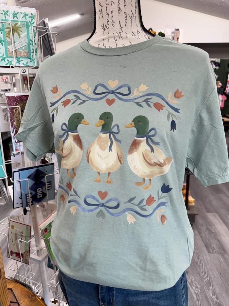 Puede incluir: Una camiseta de manga corta color verde salvia con un estampado gr&aacute;fico de tres patos &aacute;nades reales. Cada pato lleva un lazo azul alrededor del cuello. El dise&ntilde;o est&aacute; enmarcado por lazos azules, corazones peque&ntilde;os y elementos florales en tonos naranja y azul.