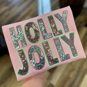 May include: A pale pink sweatshirt featuring the words "HOLLY JOLLY". The letters are embroidered with silver and burgundy sequins, outlined in green. A festive design for the holiday season.