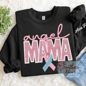 May include: Black sweatshirt featuring the text "angel MAMA" in pink glitter. A pink and blue ribbon is below the text. The sleeve has the name "Ava Faye" in white script.