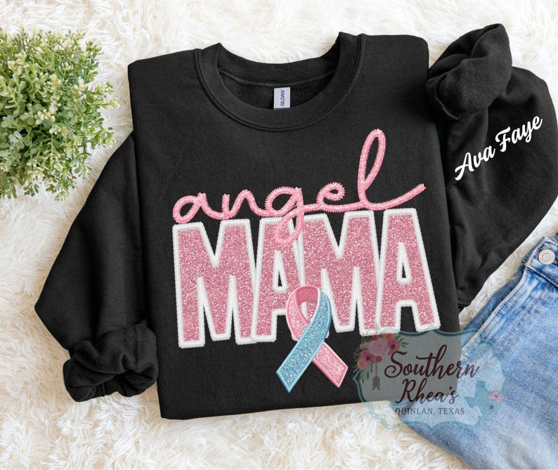 May include: Black sweatshirt featuring the text "angel MAMA" in pink glitter. A pink and blue ribbon is below the text. The sleeve has the name "Ava Faye" in white script.