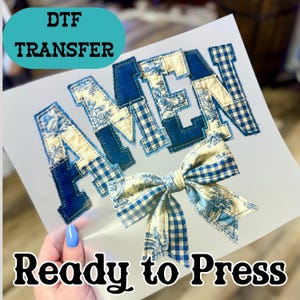 May include: A DTF transfer sheet with the word "AMEN" in a patchwork design. The letters are a mix of blue denim, floral, and gingham patterns. A matching bow is at the bottom. The text "Ready to Press" is at the bottom.