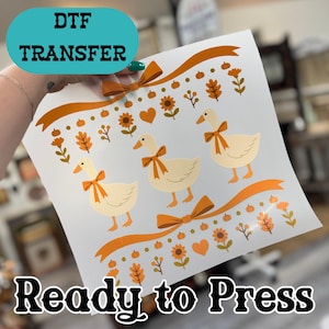 May include: A DTF transfer sheet featuring three cream-colored geese wearing orange bows, surrounded by autumn-themed icons including orange ribbons, pumpkins, sunflowers, hearts, and fall leaves on a white background with text DTF TRANSFER.