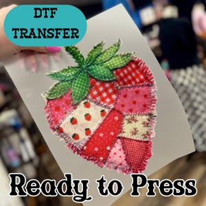 May include: DTF transfer of a strawberry design, ready to press. The strawberry is composed of various fabric patches in red, pink, and white, with a green gingham leaf. The text "Ready to Press" is below the strawberry.