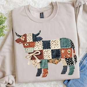 May include: Beige sweatshirt featuring a patchwork cow design. The cow is made of various fabric squares in shades of brown, blue, green, and cream, with a cream-colored bow. The sweatshirt has a crew neck and long sleeves.