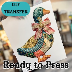 May include: A DTF transfer sheet featuring a duck graphic with a sequined, multi-colored patchwork design. The duck wears a large, layered lace and sequin bow with a central pearl-like accent. The text DTF TRANSFER and Ready to Press is visible.