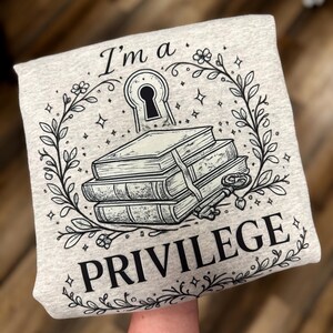 May include: A light gray sweatshirt with a black graphic design. The design includes a keyhole, a stack of books, a key, and the words "I'm a PRIVILEGE" in a decorative font. The design is surrounded by a floral wreath.