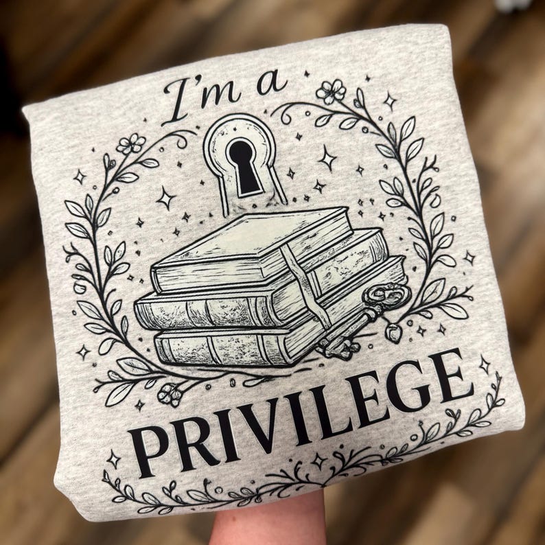 May include: A light gray sweatshirt with a black graphic design. The design includes a keyhole, a stack of books, a key, and the words "I'm a PRIVILEGE" in a decorative font. The design is surrounded by a floral wreath.