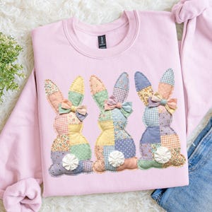 May include: A pale pink sweatshirt decorated with three appliqued patchwork bunny rabbits. Each bunny has different patterned ears, bows, and bodies, with a white pom-pom tail. The sweatshirt is made of a soft material.