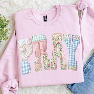 May include: A pale pink sweatshirt featuring the word "PRAY" in patchwork letters. The letters are crafted from floral and gingham fabrics. The sweatshirt is made of a soft material, perfect for casual wear.