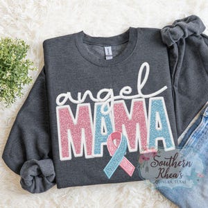 May include: A dark grey sweatshirt featuring the words "angel MAMA" in white, pink, and blue glitter lettering. A pink and blue ribbon is below the word "MAMA". The sweatshirt is displayed on a white, fluffy surface.
