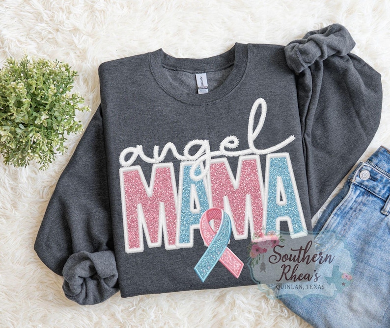 May include: A dark grey sweatshirt featuring the words "angel MAMA" in white, pink, and blue glitter lettering. A pink and blue ribbon is below the word "MAMA". The sweatshirt is displayed on a white, fluffy surface.