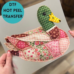 May include: A colorful DTF hot peel transfer featuring a patchwork duck design. The duck has a green checkered head, a yellow beak, and a body composed of various patterned fabrics in pink, brown, and white. The transfer is held by two hands.