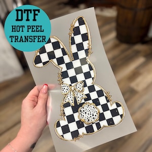 May include: A DTF (Direct to Film) hot peel transfer featuring a bunny design. The bunny is black and white checkered with gold trim and a bow. The design includes a small, dotted cloud shape.