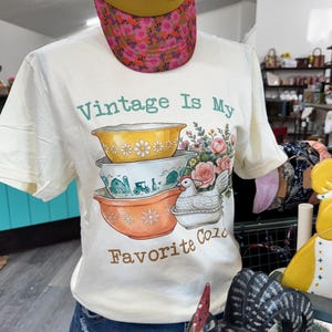 May include: A cream-colored short-sleeve t-shirt featuring a graphic of three stacked mixing bowls, a white hen, and flowers. The text reads "Vintage Is My Favorite Color" in green and brown. The shirt is worn with a yellow and floral print cap.