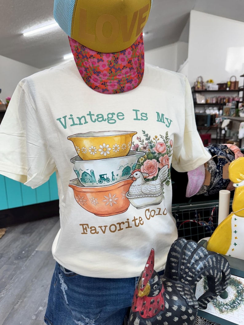 May include: A cream-colored short-sleeve t-shirt featuring a graphic of three stacked mixing bowls, a white hen, and flowers. The text reads "Vintage Is My Favorite Color" in green and brown. The shirt is worn with a yellow and floral print cap.