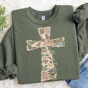 May include: An olive green sweatshirt with a camouflage cross design. The cross is made from patches of green, brown, and tan fabrics. The words "He is Risen" are written in white script on the right side of the cross.