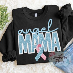 May include: Black crew neck sweatshirt featuring the words "angel MAMA" in light blue glitter lettering. A light blue and pink ribbon is below the word "MAMA". The sweatshirt is on a white, textured surface.