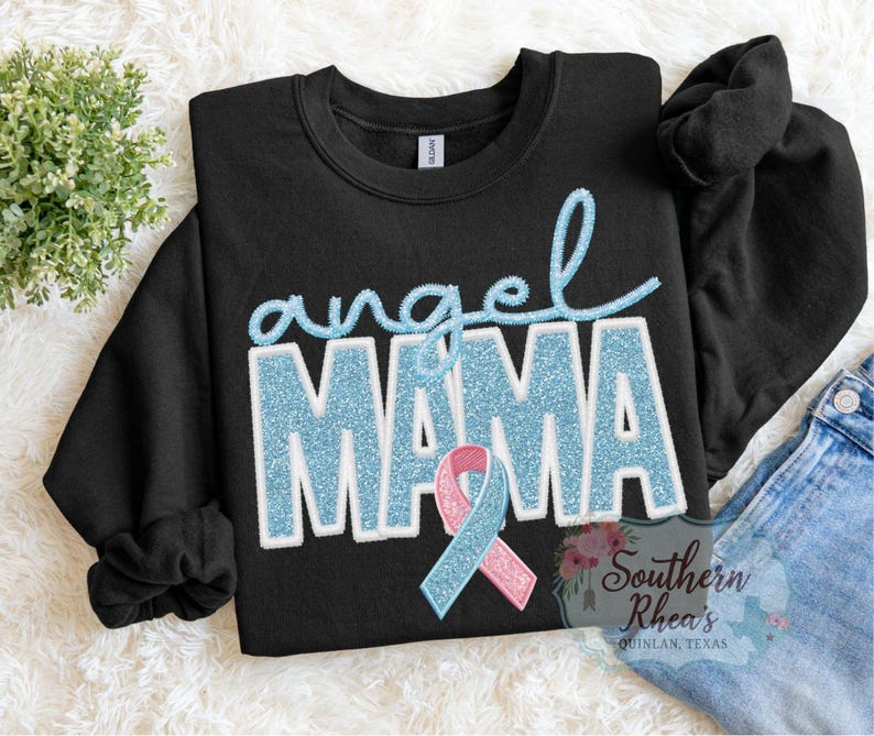 May include: Black crew neck sweatshirt featuring the words "angel MAMA" in light blue glitter lettering. A light blue and pink ribbon is below the word "MAMA". The sweatshirt is on a white, textured surface.
