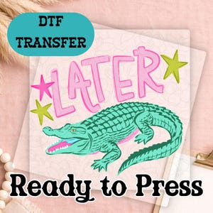 May include: A DTF transfer with a teal alligator illustration and the word "LATER" in pink. The phrase "Ready to Press" is printed in black below the alligator design. Pink and green star accents complete the design.