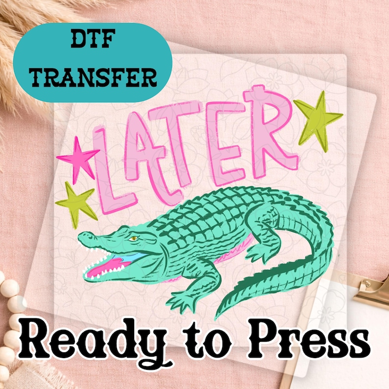 May include: A DTF transfer with a teal alligator illustration and the word "LATER" in pink. The phrase "Ready to Press" is printed in black below the alligator design. Pink and green star accents complete the design.