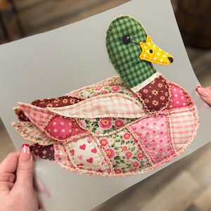 May include: A handmade patchwork duck with a green checkered head and a yellow beak. The duck's body features a variety of pink and brown floral and heart-patterned fabrics. The duck is held against a light gray background.