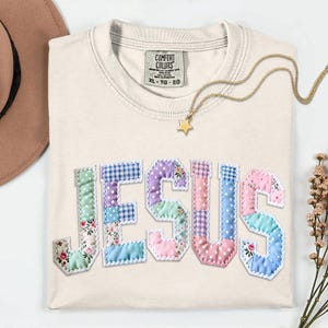 May include: Cream-colored t-shirt with the word "JESUS" appliqued in patchwork fabric. The letters feature floral, polka dot, and gingham patterns in pastel colors. A gold star pendant necklace and a Comfort Colors tag are visible.