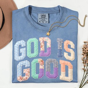 May include: A blue t-shirt with the text GOD IS GOOD in large, multi-patterned fabric letters featuring floral, gingham, and polka dot prints. The shirt has a Comfort Colors label in the neck and is paired with a gold-toned star pendant necklace.