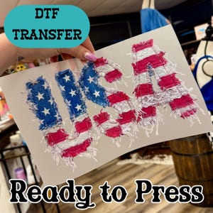 May include: A DTF (Direct to Film) transfer featuring the word "USA" in a distressed, patriotic design. The letters are made of red, white, and blue fabric, with a frayed edge effect. The text "Ready to Press" is also visible.