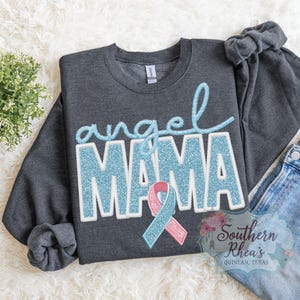 May include: A charcoal grey sweatshirt featuring the words "angel MAMA" in light blue glitter lettering and a pink and blue ribbon. The sweatshirt is displayed on a white fluffy surface with a small plant and a pair of blue jeans.