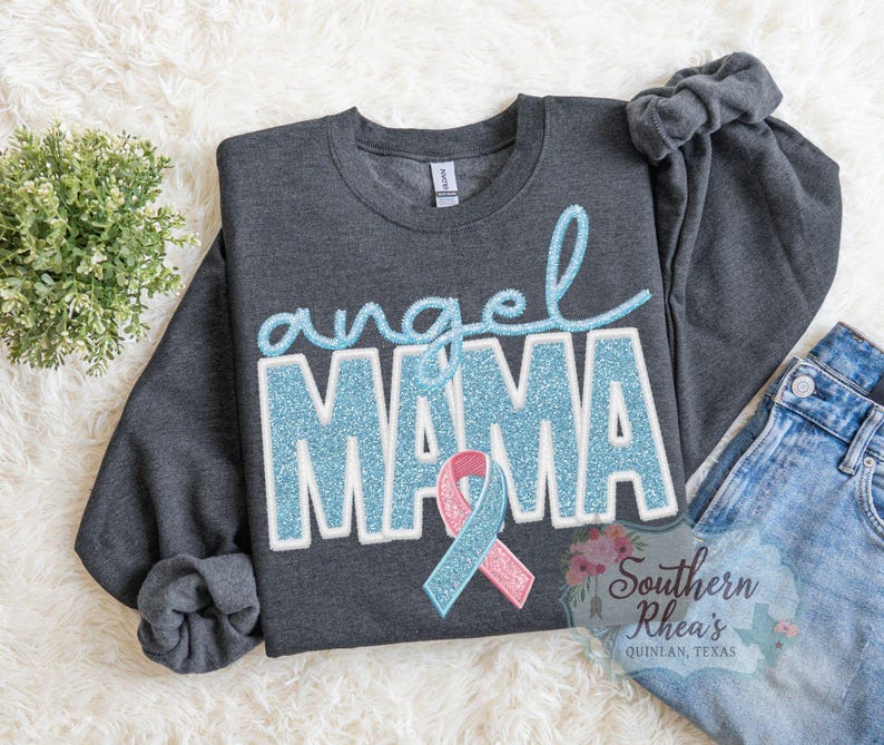 May include: A charcoal grey sweatshirt featuring the words "angel MAMA" in light blue glitter lettering and a pink and blue ribbon. The sweatshirt is displayed on a white fluffy surface with a small plant and a pair of blue jeans.