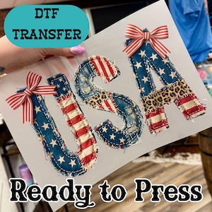 May include: DTF transfer featuring the letters "USA" in a patchwork design. The letters are adorned with red, white, and blue fabrics, including stars, stripes, and leopard print. Each letter has a red and white striped bow. The text "Ready to Press" is at the bottom.
