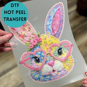 May include: A colorful DTF hot peel transfer featuring a bunny face with heart-shaped glasses. The bunny has floral patterned ears, a yellow and blue face, and pink accents. The transfer is embellished with pearls and roses.