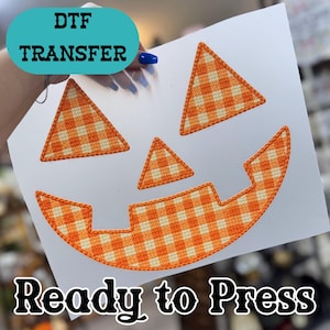 May include: A DTF transfer sheet featuring a jack-o'-lantern face design with two triangular eyes, a triangular nose, and a wide mouth, all filled with an orange and white gingham check pattern. The text DTF TRANSFER and Ready to Press appear.