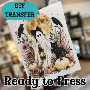 May include: A DTF transfer sheet featuring a white ghost with a black bow holding a lantern, a black cat, two black crows, a large yellow moon, pumpkins, and floral elements. The text DTF TRANSFER and Ready to Press is printed on the sheet.
