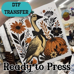 May include: A DTF transfer sheet featuring a graphic illustration of a goose wearing a black bow tie, surrounded by orange and black flowers, foliage, and a large pumpkin. The design includes text that reads DTF TRANSFER and Ready to Press.