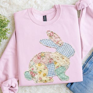 May include: A light pink sweatshirt featuring a patchwork bunny design. The bunny is made of various fabrics, including floral prints, gingham, and lace. The sweatshirt is a crew neck style.