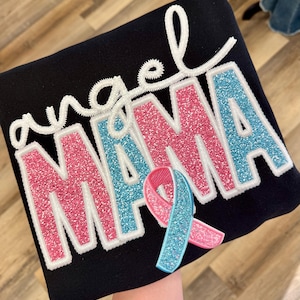 May include: Black sweatshirt with the words "angel MAMA" in pink and blue glitter lettering, outlined in white. A pink and blue ribbon is attached to the bottom of the text.
