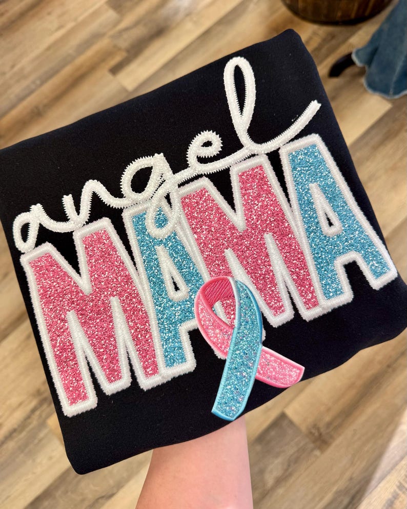 May include: Black sweatshirt with the words "angel MAMA" in pink and blue glitter lettering, outlined in white. A pink and blue ribbon is attached to the bottom of the text.