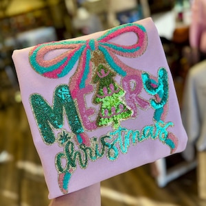 May include: A light pink sweatshirt featuring a large sequined design of a bow and a Christmas tree. The text MERRY CHRISTMAS is written in green and teal sequins. The bow is outlined in pink and teal sequins, and the tree is green and lime.