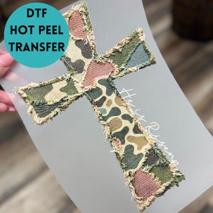 May include: A DTF hot peel transfer featuring a cross design with a camouflage pattern in shades of green, brown, and tan. The cross has a frayed edge and the words "He is Risen" are written in white.