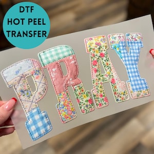 May include: DTF Hot Peel Transfer featuring the word "PRAY" in patchwork fabric letters. The letters are made of various floral and gingham patterns in pink, blue, and yellow. The letters are outlined with a decorative stitch.