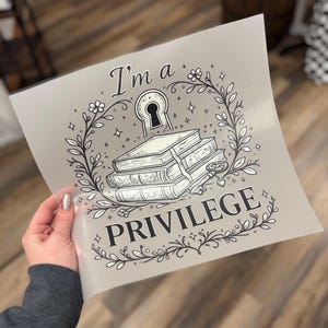 May include: A gray print featuring the text "I'm a PRIVILEGE" with a keyhole, books, and floral elements. The design is in black and white, with a decorative border of leaves and stars.