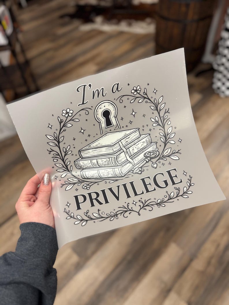 May include: A gray print featuring the text "I'm a PRIVILEGE" with a keyhole, books, and floral elements. The design is in black and white, with a decorative border of leaves and stars.