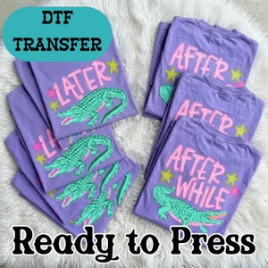 May include: A stack of purple t-shirts with a teal alligator graphic and the words "LATER" and "AFTER WHILE" in pink and yellow. The text "DTF TRANSFER" and "Ready to Press" are also visible.