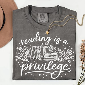 May include: Dark gray t-shirt with the text "reading is a privilege" in white script and a stack of books graphic. A gold necklace with a star pendant rests on the shirt. The shirt is folded and has a Comfort Colors tag.