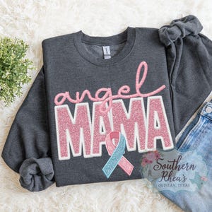 May include: Dark grey sweatshirt with the words "angel MAMA" in pink glitter lettering. A pink and blue ribbon is below the text. The sweatshirt is folded on a white, textured surface with a small green plant and a pair of blue jeans.