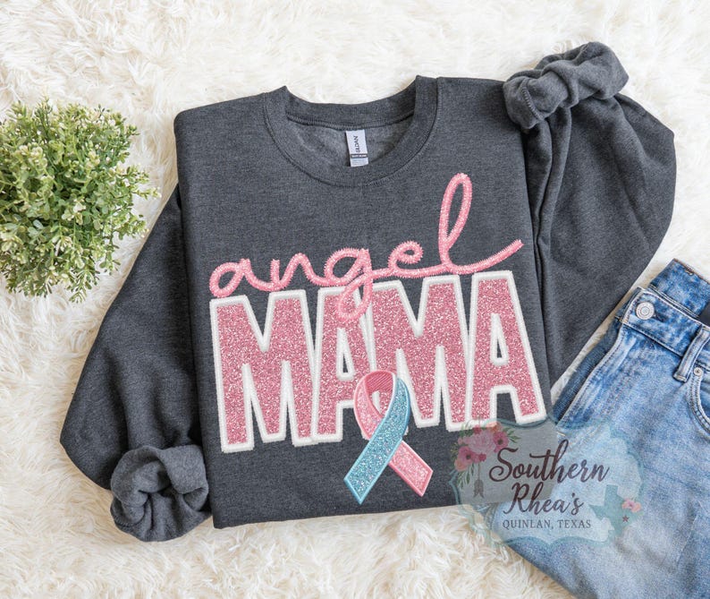 May include: Dark grey sweatshirt with the words "angel MAMA" in pink glitter lettering. A pink and blue ribbon is below the text. The sweatshirt is folded on a white, textured surface with a small green plant and a pair of blue jeans.