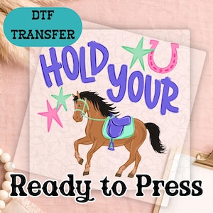 May include: DTF transfer with the text "HOLD YOUR" in purple, a brown horse with a blue saddle, and the text "Ready to Press" in black. The design includes pink and green stars and a pink horseshoe.